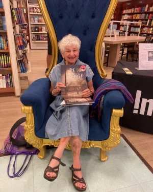 A smiling white-haired woman sits in an oversized blue chair holding a children's book